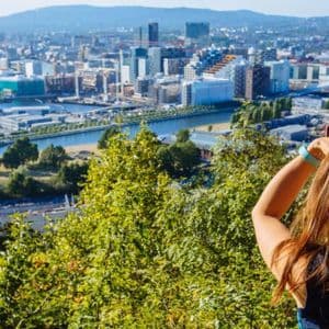 Una donna con uno zaino, vista da dietro, fotografa un moderno paesaggio urbano con un fiume da un punto di vista elevato.