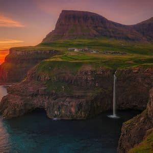 Una cascata si getta da una scogliera erbosa nell'oceano al tramonto, con un piccolo villaggio sulla cima della scogliera.