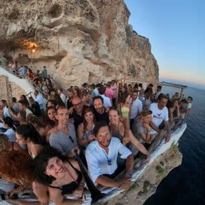 Eine WeRoad Gruppenreise macht ein Weitwinkel-Selfie auf einer belebten Bartrasse, die in einen Felsen über dem Meer gebaut ist.