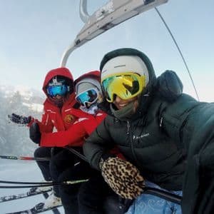 Drei Personen einer WeRoad-Gruppe in Skiausrüstung machen ein Selfie auf einem Skilift vor verschneiter Bergkulisse.