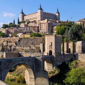 Un grande ponte in pietra con archi, che attraversa un fiume, porta a una storica città fortificata su una collina sotto un cielo azzurro e sereno.