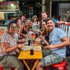 Un gruppo WeRoad sorride mentre mangia noodles con le bacchette a un tavolo di legno accanto a una bancarella di street food.