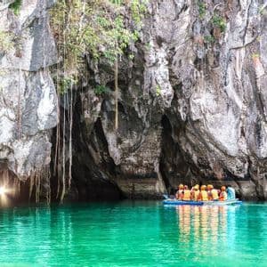 Un viaggio di gruppo WeRoad in barca su acqua turchese si avvicina all'ingresso illuminato dal sole di una grotta marina in una scogliera rocciosa.