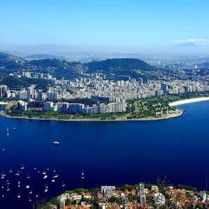 Vue aérienne d'une ville côtière le long d'une baie remplie de voiliers, nichée contre des collines verdoyantes sous un ciel bleu clair.