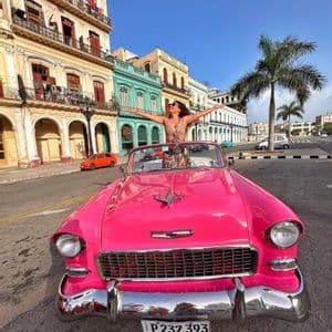 Eine Frau steht mit ausgestreckten Armen in einem knallrosa Vintage-Cabrio auf einer Stadtstraße, gesäumt von bunten Gebäuden und einer Palme.