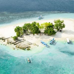 Veduta aerea di una piccola isola tropicale con spiaggia di sabbia bianca, alberi verdi e diverse imbarcazioni in acqua turchese.