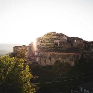 Un borgo di vecchie case costruite su un fianco di collina verdeggiante, con il sole nascente che crea un bagliore luminoso da dietro gli edifici.