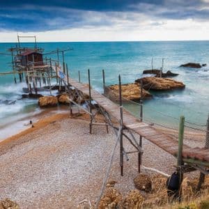 Un lungo molo di legno conduce da una spiaggia di ciottoli a una capanna di pescatori su palafitte nel mare turchese sotto un cielo nuvoloso.