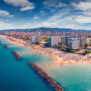 Vista aerea di una lunga spiaggia sabbiosa affollata di ombrelloni e persone, accanto a una città con colline sullo sfondo.
