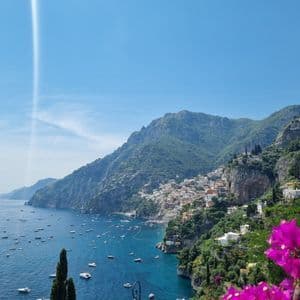 Un pueblo costero encaramado en una ladera verde y empinada, con vistas a una bahía azul repleta de barcos y flores rosas en primer plano.