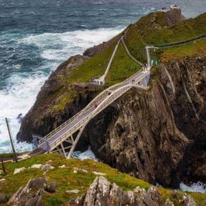 Eine moderne Fußgängerbrücke überquert eine Schlucht zwischen zwei steilen, grasbewachsenen Klippen, während Menschen darüber über einem stürmischen Meer gehen.
