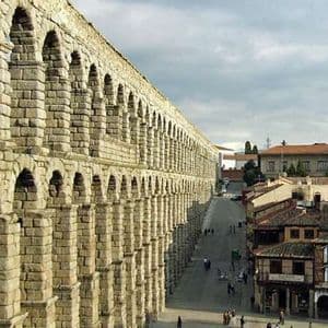 Un antico acquedotto in pietra a due livelli si estende attraverso una storica piazza cittadina con persone che camminano sotto.