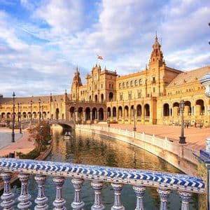 An ornate building with a canal is seen from a bridge with a decorative blue and white tiled balustrade under a partly cloudy sky.