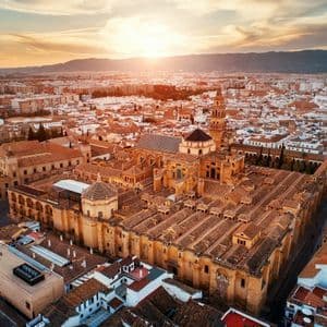 An aerial view of a sprawling city with terracotta roofs centered around a large historic cathedral complex at sunset.