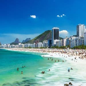 A crowded city beach with people swimming in turquoise water and relaxing on white sand, with mountains in the background.