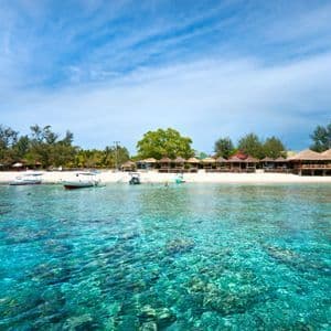 Acqua turchese cristallina con una barriera corallina visibile davanti a una spiaggia di sabbia bianca costeggiata da bungalow con tetto di paglia e barche.