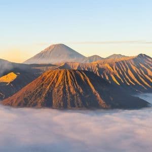 Un volcan fumant et d'autres sommets montagneux s'élèvent au-dessus d'une dense couche de nuages, illuminés par la lumière dorée du lever du soleil.