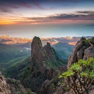 Una vista panoramica da una montagna rocciosa su una valle verde e una città costiera illuminata sul mare al tramonto.