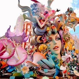 Una scultura imponente e colorata, raffigurante un grande volto femminile, figure fantastiche e fiori, si erge all'aperto in una città.