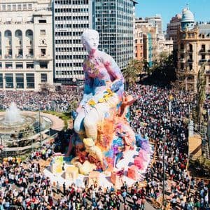 Una grande statua colorata di una figura seduta in una piazza cittadina, circondata da una folla immensa durante un festival soleggiato.