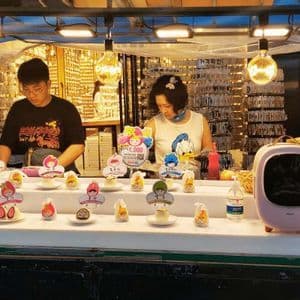 Un uomo e una donna lavorano di notte a una bancarella di street food, preparando piccoli dolci di frutta esposti su un bancone bianco sotto luci calde.