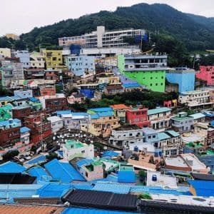 Un denso grappolo di case colorate con tetti blu brillanti costruite su un rigoglioso fianco di collina verde sotto un cielo nuvoloso.