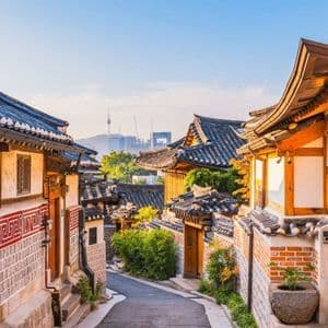 Case tradizionali coreane con tetti a tegola fiancheggiano una strada stretta, con lo skyline di una città moderna e una torre sullo sfondo.
