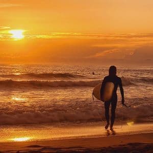Un surfista in muta trasporta una tavola da surf e cammina lungo la spiaggia verso le onde durante un tramonto arancione splendente.