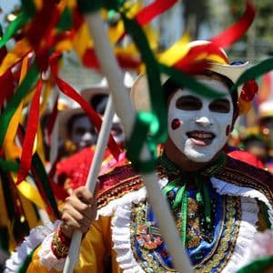Una persona in costume colorato e con il volto dipinto di bianco sorride durante una parata, tenendo un bastone decorato con nastri.