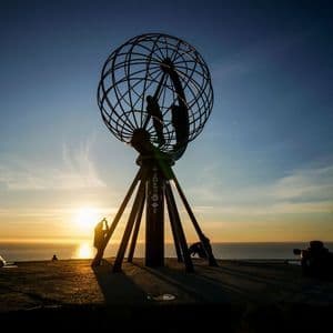 Un voyage de groupe WeRoad en silhouette devant un coucher de soleil, près d'une grande sculpture de globe dominant l'océan.