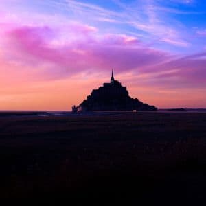 Eine Burg auf einer Insel, die sich vor einem lebhaften Himmel mit rosa und violetten Wolken im Sonnenuntergang abzeichnet.