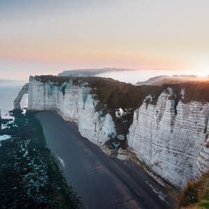 Acantilados de tiza blanca, incluyendo un arco natural y un farallón, se elevan sobre una playa oscura y un mar en calma al amanecer.