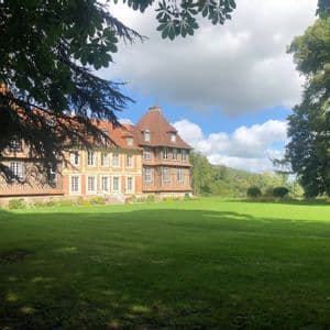 Un grand manoir à colombages avec un toit de tuiles rouges se dresse à côté d'une vaste pelouse verte, encadré par des branches d'arbres feuillus.