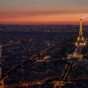 Una vista aerea di un paesaggio urbano al crepuscolo, con la Torre Eiffel illuminata che proietta un fascio di luce sulle luci della città.