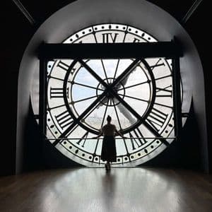 Una donna in silhouette si trova su un pavimento di legno, guardando un panorama urbano attraverso una grande finestra a orologio.