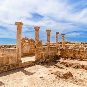 Diverse colonne di pietra consumate dal tempo si ergono tra antiche rovine in un sito archeologico sotto un cielo azzurro parzialmente nuvoloso.