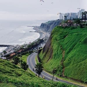 Parapendisti sorvolano una tortuosa strada costiera e verdi scogliere a picco sull'oceano, con edifici cittadini sullo sfondo.