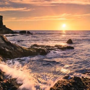 Meereswellen brechen sich an einer felsigen Küste unter einem goldenen Sonnenuntergang, während die Sonne tief am Horizont steht.