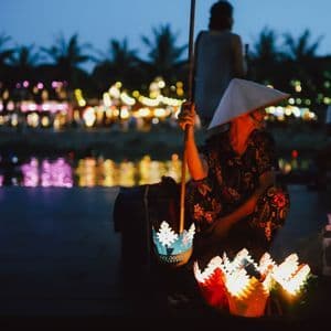 Un venditore con un cappello conico vende lanterne di carta colorate e illuminate sulla riva di un fiume al tramonto, con le luci della città sullo sfondo.
