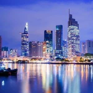 Uno skyline moderno con grattacieli illuminati di notte, che si riflette su un fiume con barche ormeggiate in primo piano.