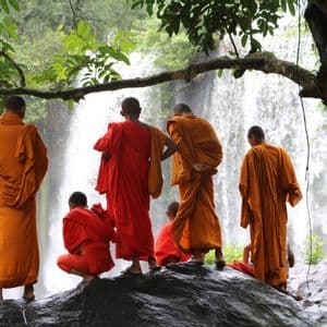 Un gruppo di monaci buddisti con tuniche arancioni e rosse si trova su una roccia bagnata, osservando una grande cascata in una foresta lussureggiante.