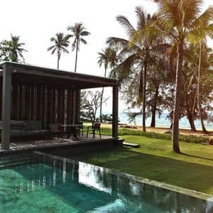 Una piscina e una cabana in legno su un prato erboso, accanto a una spiaggia sabbiosa con palme e l'oceano.