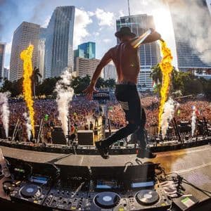 Un artista a torso nudo con un cappello salta su un palco con fuochi d'artificio di fronte a una folla immensa a un festival musicale all'aperto.