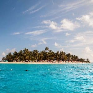 Un'isola tropicale con palme e tende colorate su una spiaggia sabbiosa, vista dal mare turchese sotto un cielo parzialmente nuvoloso.