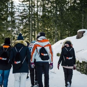 Rear view of a WeRoad group trip walking along a snowy path through a pine forest, wearing winter jackets and backpacks.