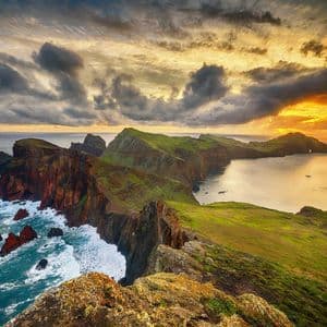 Una lunga penisola verde, con ripide scogliere rocciose, si protende nell'oceano sotto un cielo nuvoloso al tramonto.