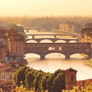 Vue aérienne d'une ville historique avec des toits en terre cuite et une rivière traversée par plusieurs ponts au coucher du soleil doré.