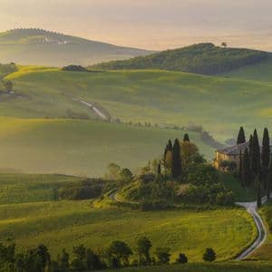 Une villa entourée de cyprès se dresse sur une colline verdoyante dans un paysage brumeux au lever du soleil.