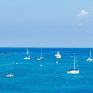 Vista panoramica di diverse barche a vela su un mare blu intenso, accanto a una grande scogliera rocciosa, sotto un cielo limpido.