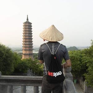 Una persona vista de espaldas, con un sombrero cónico y una mochila WeRoad, contempla una alta pagoda desde un mirador de piedra.